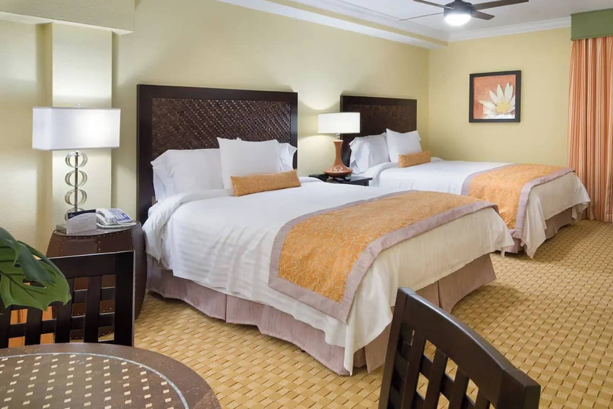 A hotel room with two beds, a desk, a chair, a lamp, a telephone, a picture on the wall, and a window with curtains.