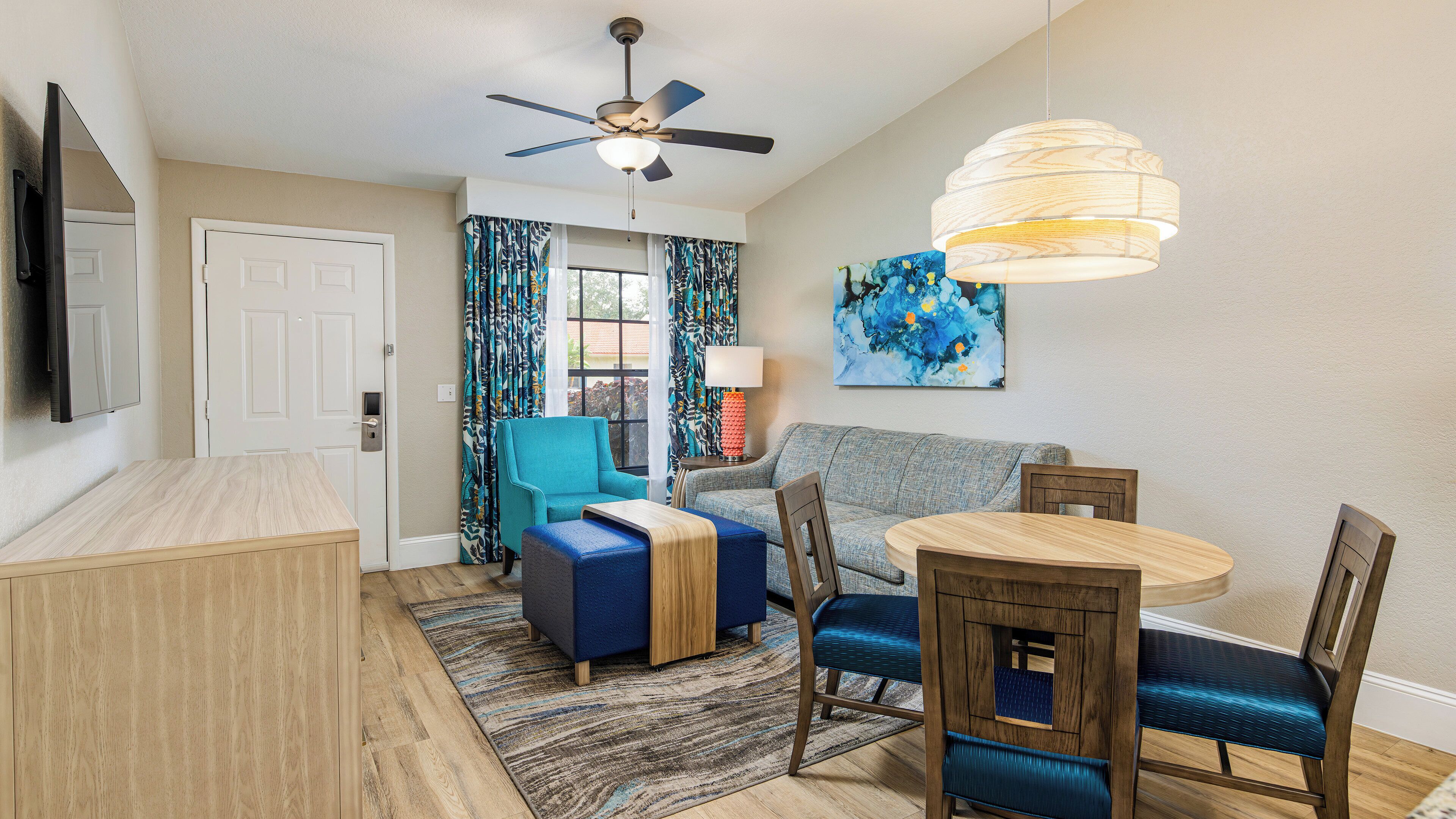 A modern living room with a ceiling fan, a dining table with chairs, a sofa, a TV mounted on the wall, and a painting.