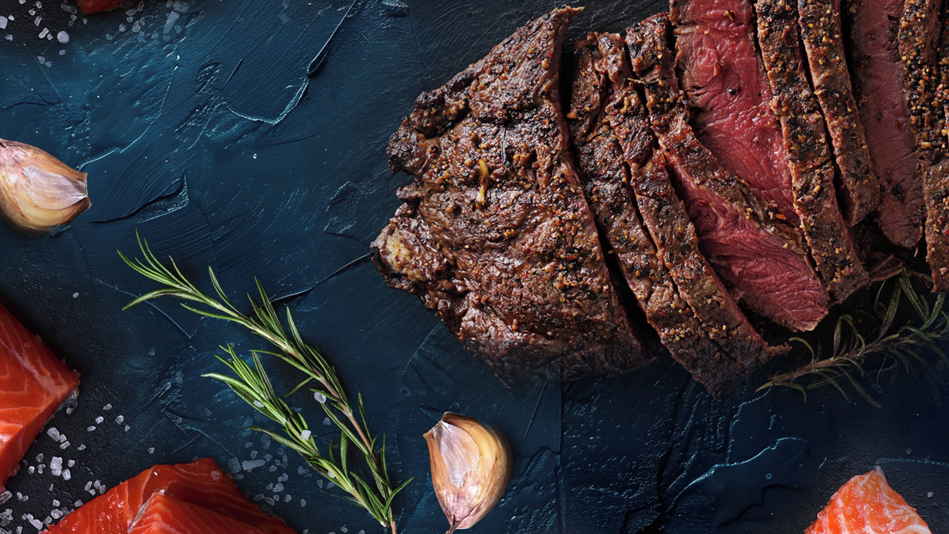 Een steak met kruiden, omringd door teentjes knoflook en rozemarijn op een donkere ondergrond.