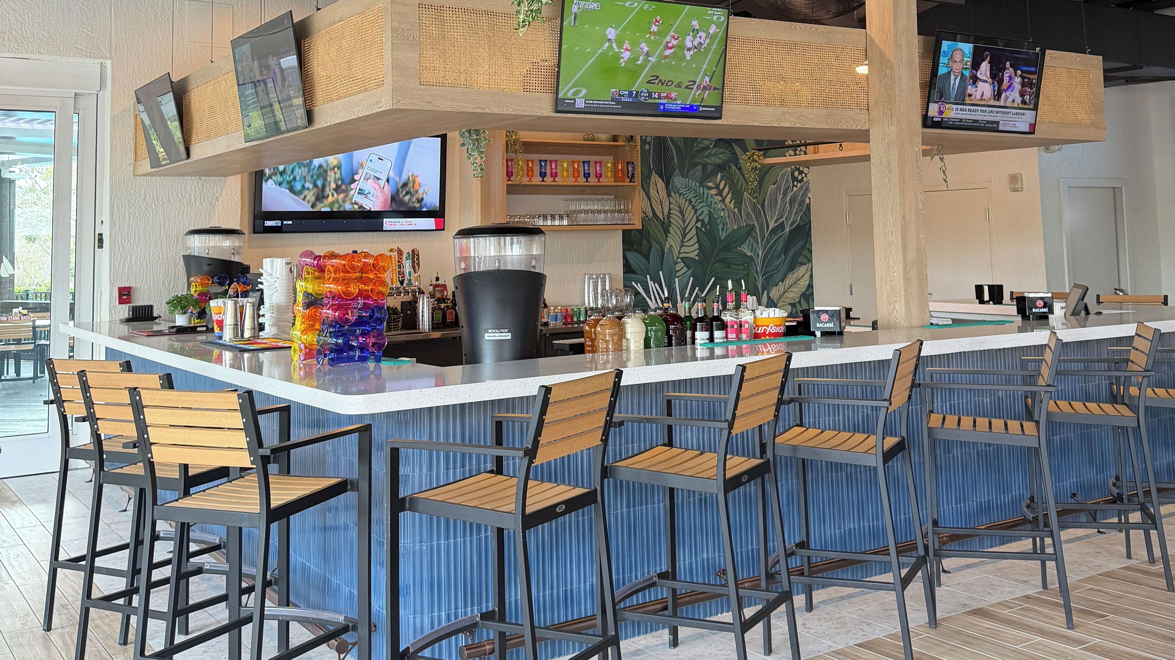 A bar area with multiple seating options, a counter with various beverages, and screens displaying sports events.