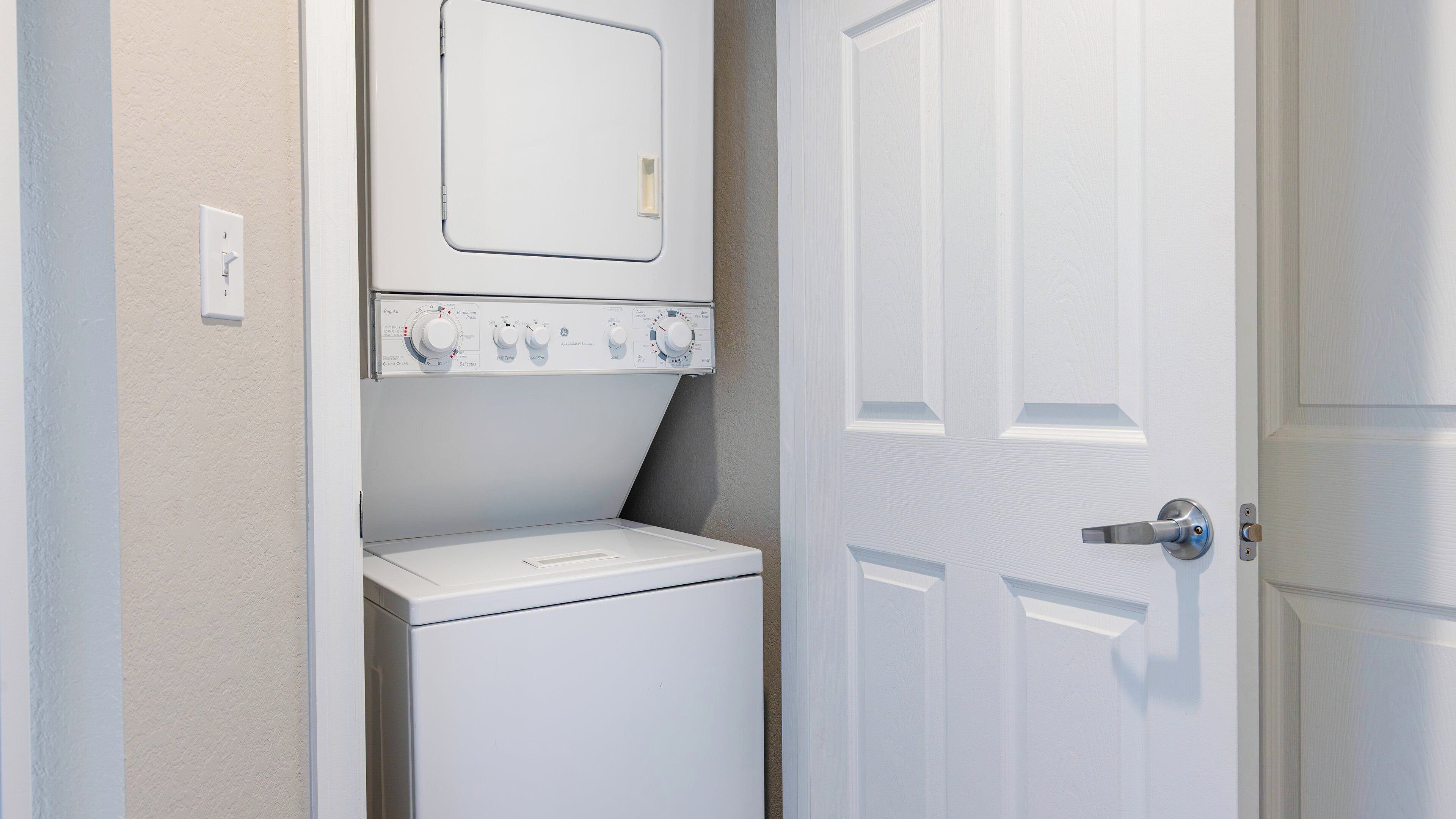 A laundry room with a front-loading washer and dryer, a door with a silver handle, and a light switch on the wall.