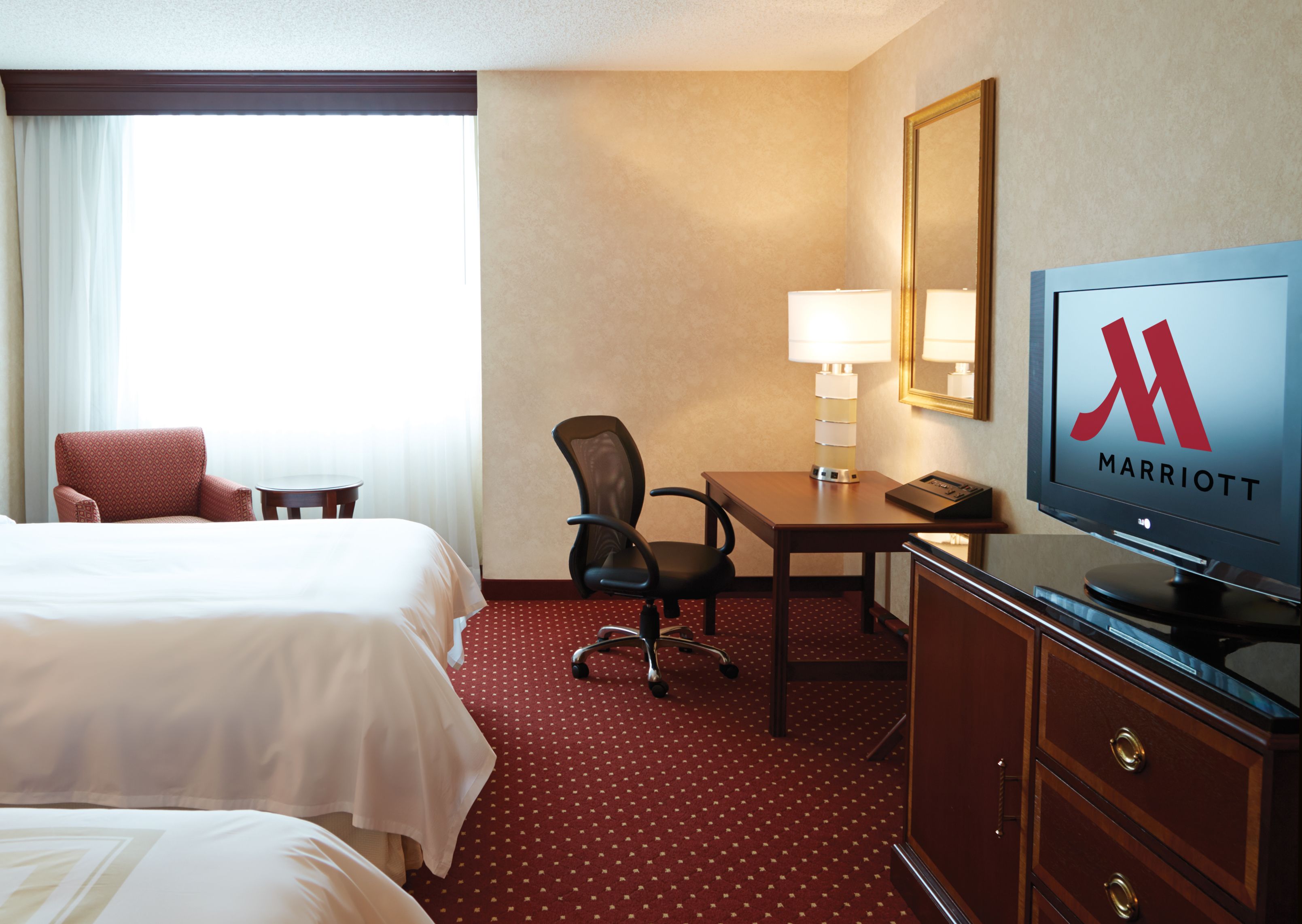 Habitación de hotel con dos camas, un escritorio con lámpara, una silla, un televisor que muestra el logotipo de Marriott y un espejo grande en la pared.