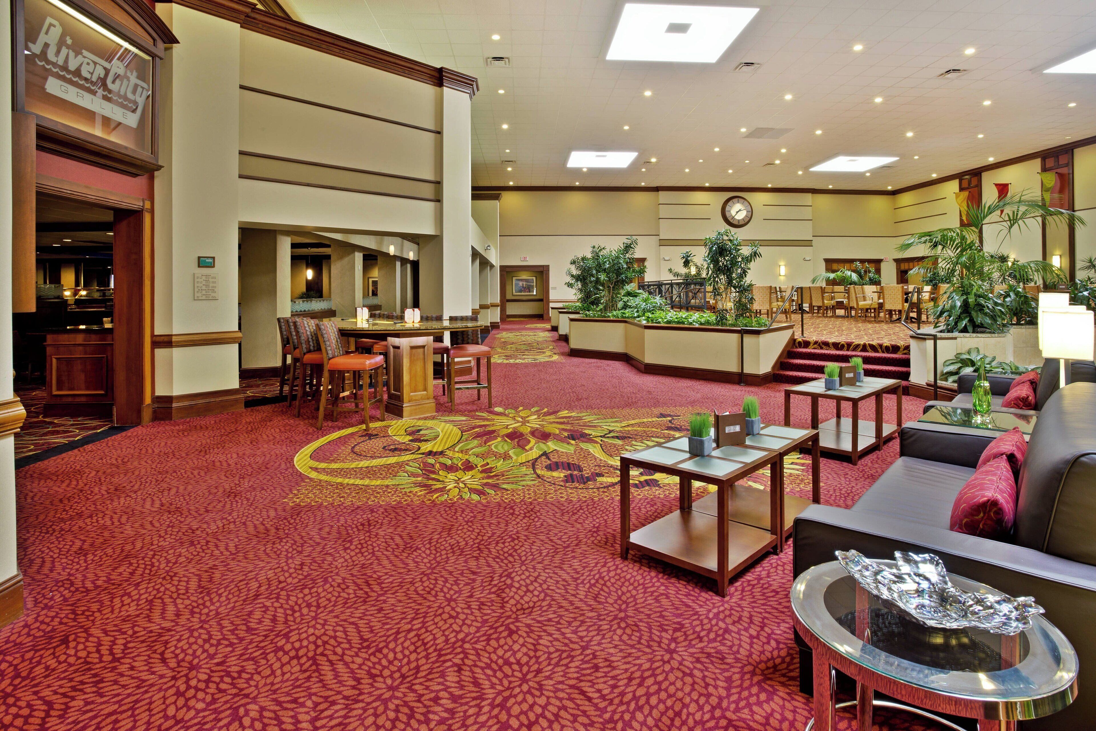 Un hall de hotel con alfombra roja, zonas de estar, un bar y una escalera que sube a otro nivel.