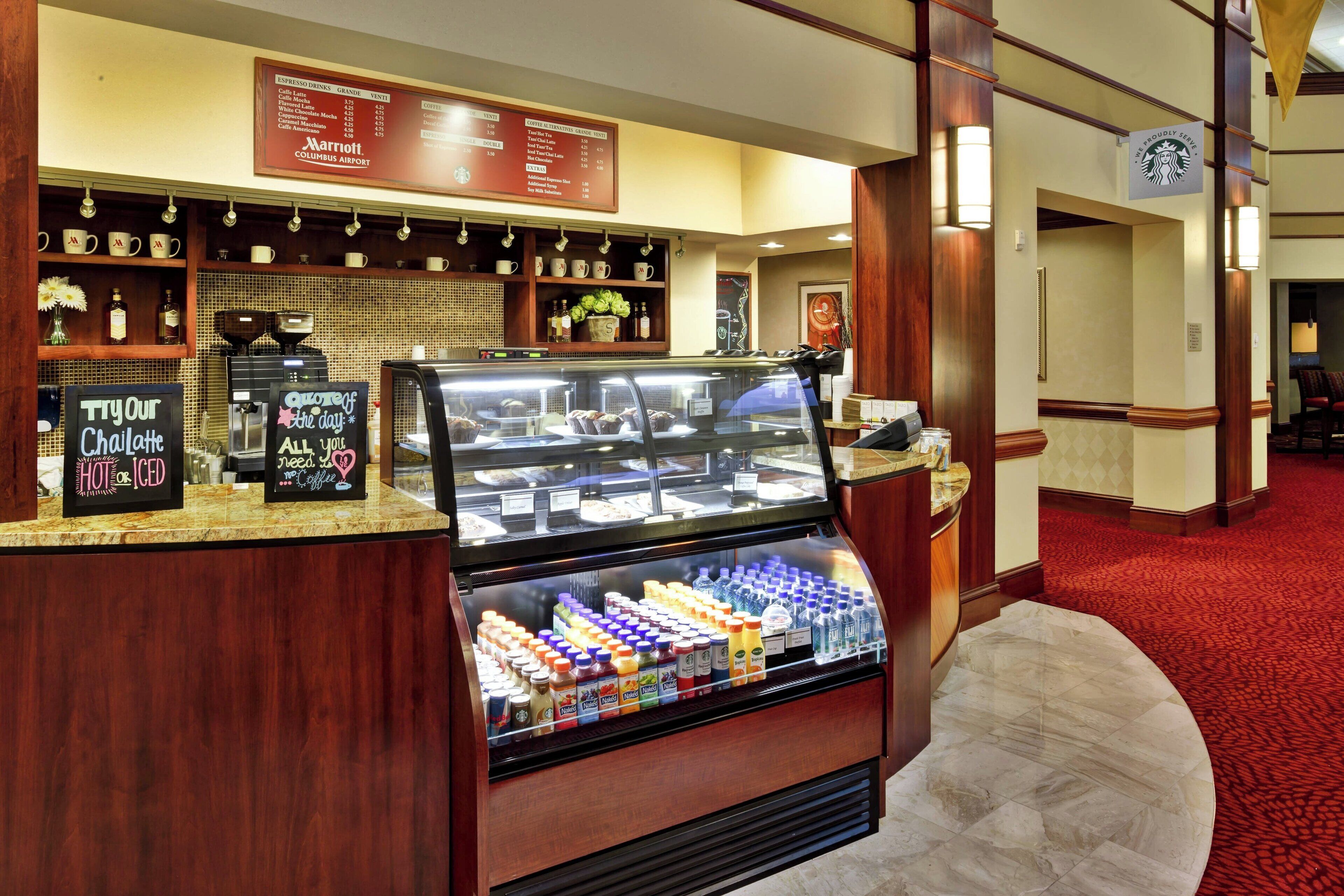 Cafetería de hotel con un menú, máquinas de café y una vitrina con bebidas embotelladas.