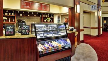 Cafetería de hotel con un menú, máquinas de café y una vitrina con bebidas embotelladas.