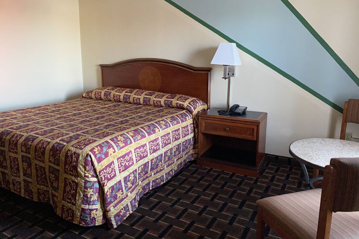 A hotel room with a bed, a nightstand with a lamp, a chair, and a patterned carpet.