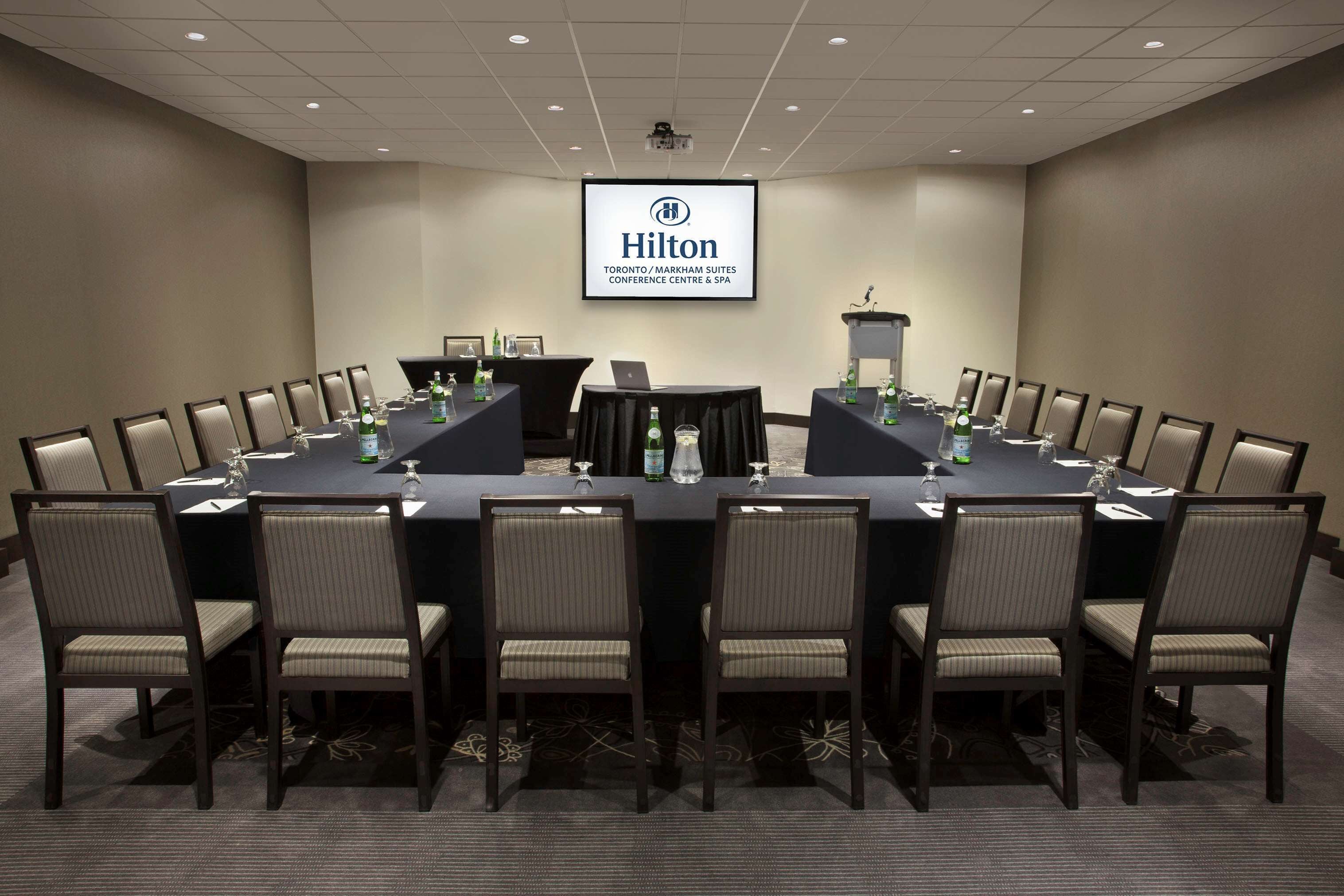 Una sala conferenze con un ampio tavolo, sedie e uno schermo proiettore che mostra il logo Hilton.
