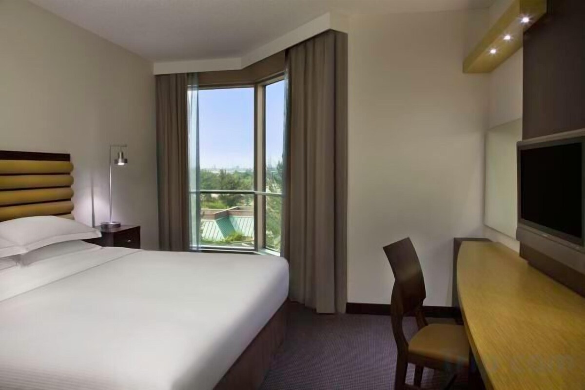 Ein Hotelzimmer mit einem großen Bett, einem Schreibtisch, einem Sessel, einem Fernseher und einem Balkon mit Vorhängen.