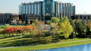 A modern hotel with a central clock tower, surrounded by a well-maintained park with trees and a body of water. - Hilton Toronto/Markham Suites Conference Centre & Spa (Markham)