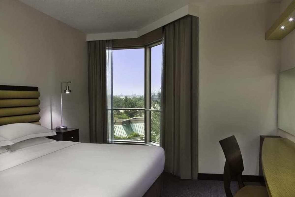 A hotel room with a large bed, a desk, a chair, a nightstand, a lamp, and a view of the outdoors through a window.
