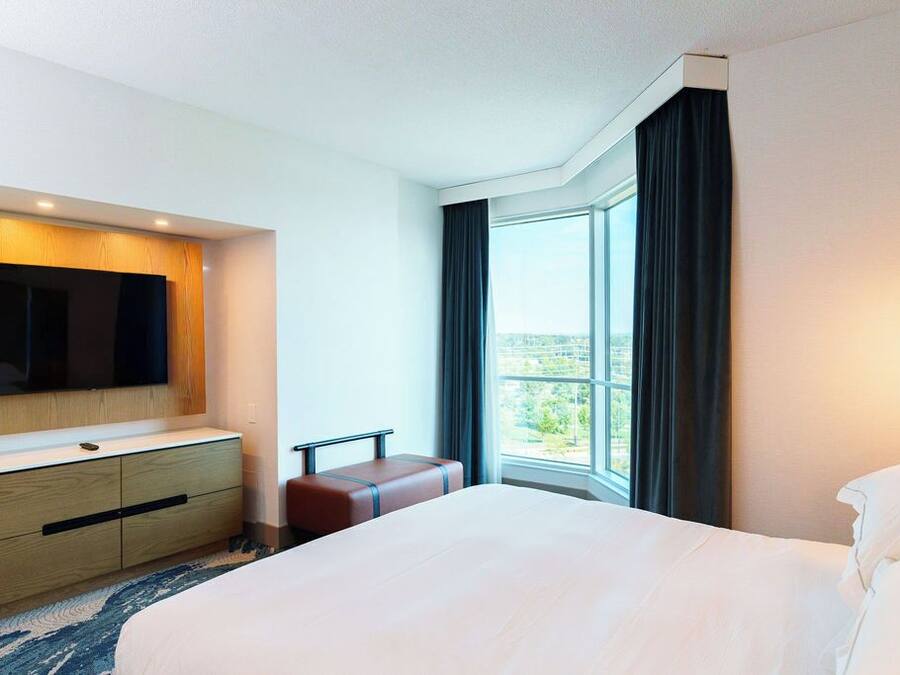 Habitación de hotel con una cama grande, un televisor de pantalla plana montado en un panel de madera, una pequeña zona de estar y un amplio ventanal con vistas al exterior.