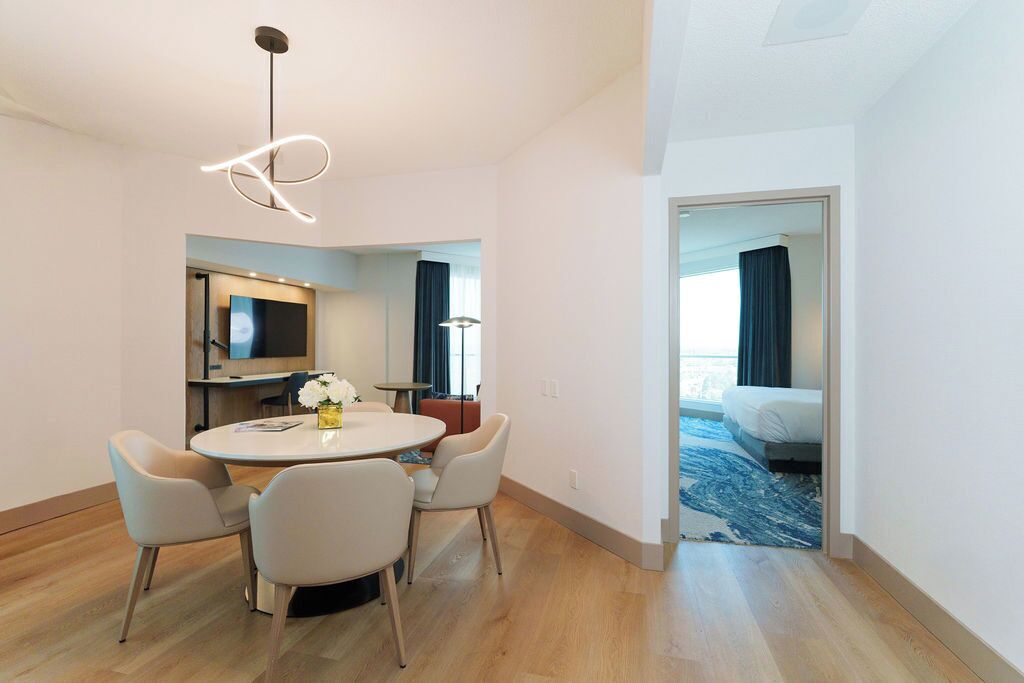 A modern hotel room with a round dining table, four chairs, a TV, and a bed with a blue carpet.