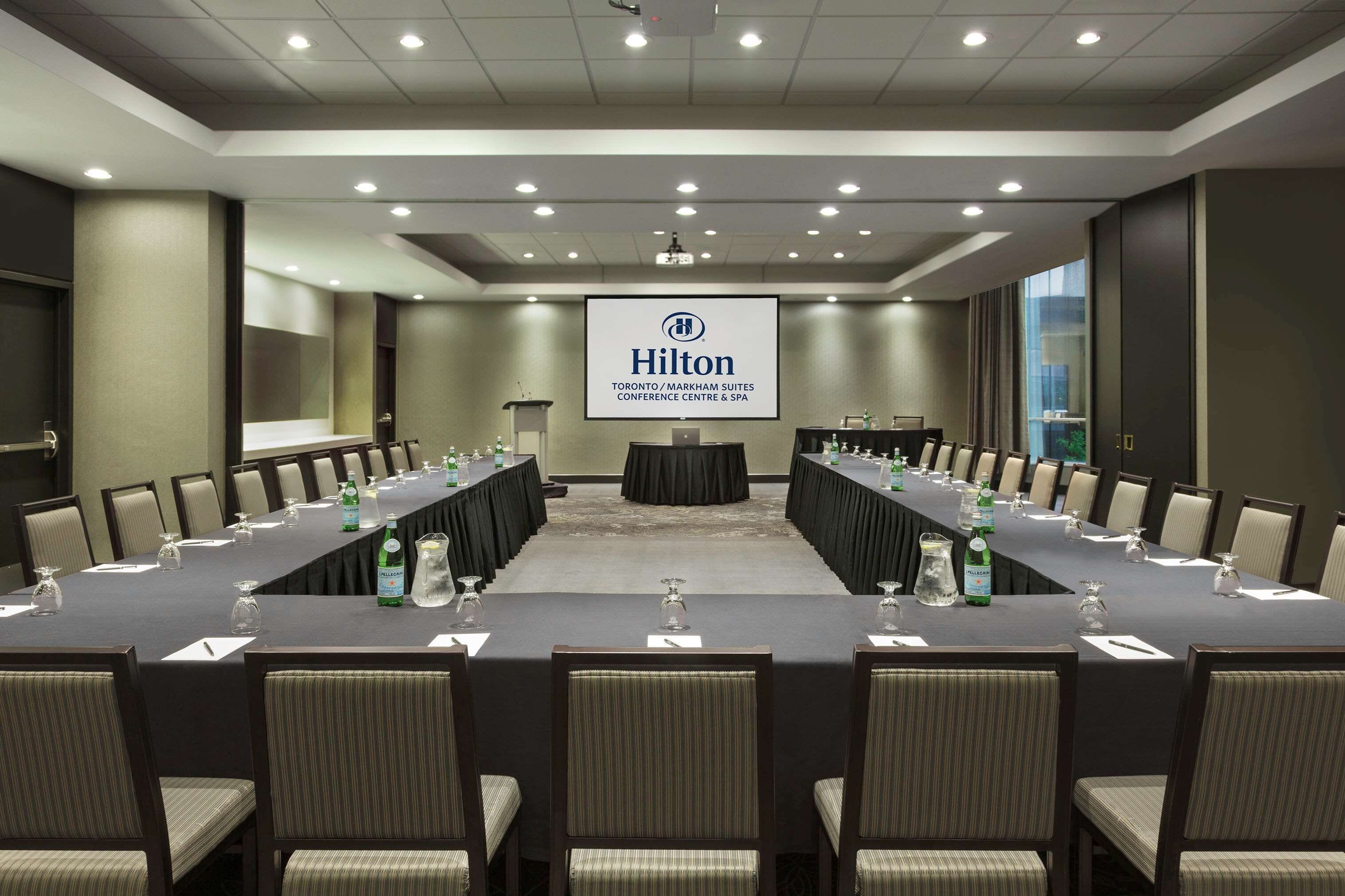 Un salón de conferencias con una larga mesa, sillas y una pantalla que muestra el logotipo de Hilton.