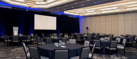 Un salón de conferencias con mesas redondas, sillas, una pantalla de proyector y un atril.