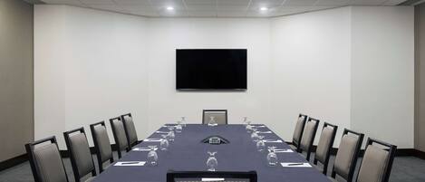 Un salón de conferencias con una mesa larga, sillas, un televisor de pantalla plana y un reloj en la pared.