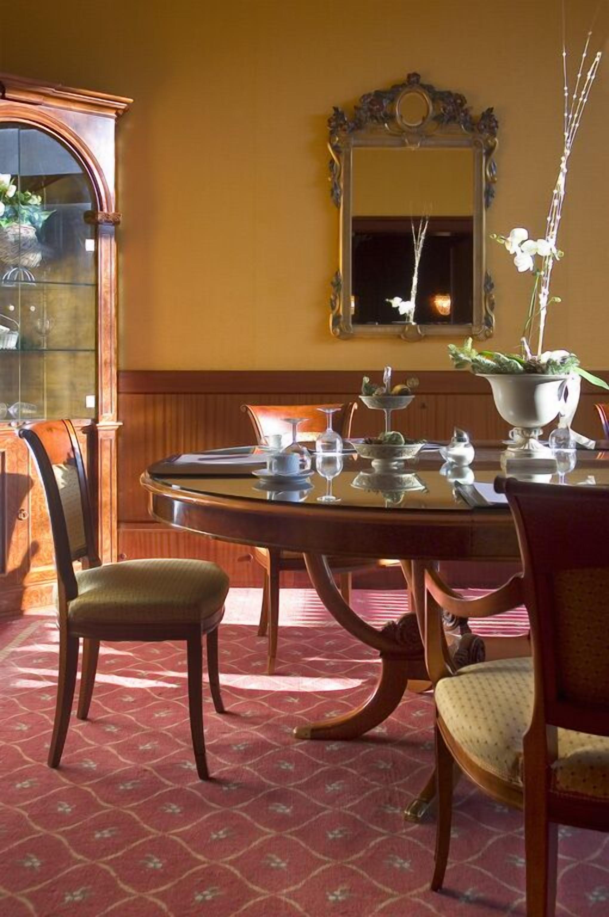 A dining area with a round table set for four, surrounded by chairs. A wooden cabinet with glass doors is to the left, and a mirror with an ornate frame hangs on the wall above the table.
