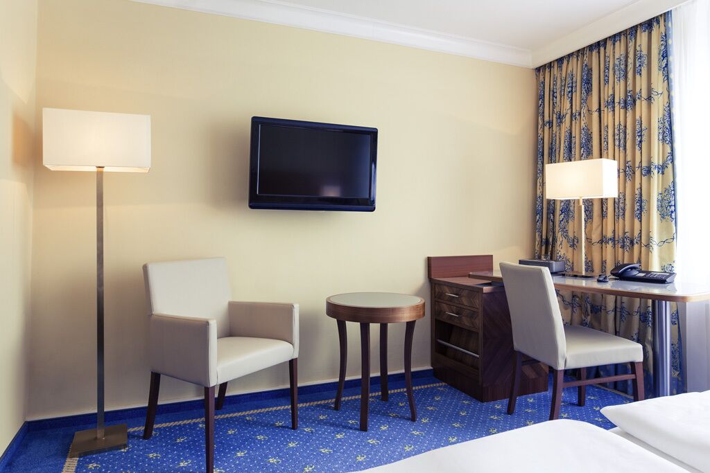 Een hotelkamer met een bed, twee stoelen, een bureau met een computer, een aan de muur bevestigde televisie en een kamer met een patroon.