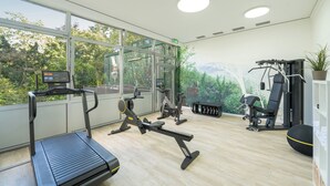 Een moderne sportschool met loopband, elliptische trainer en gewichtstoestellen, grote ramen met uitzicht op bomen en bergen, en een houten vloer.