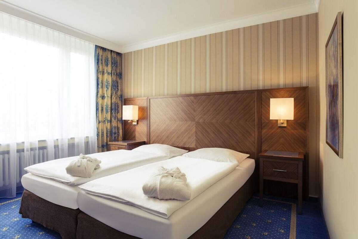 Ein Hotelzimmer mit zwei Betten, einem hölzernen Kopfteil, Nachttischlampen, einem blauen Teppich und einem Fenster mit transparenten Vorhängen.