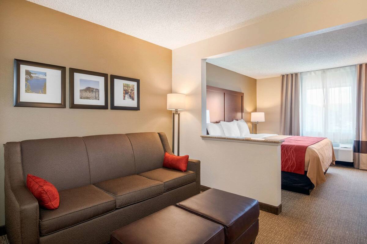 A hotel room with a sofa, a small table, a bed with a red blanket, a mirror, and three framed pictures on the wall.