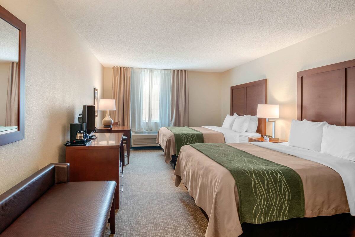 A hotel room with two beds, a desk with a lamp, a chair, and a large window with curtains.