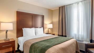A hotel room with a large bed, two bedside tables with lamps, a window with curtains, and a wall-mounted air conditioning unit.
