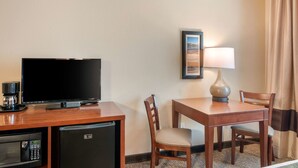 A hotel room with a flat-screen TV, a coffee maker, a refrigerator, a table with two chairs, a lamp, and a framed picture on the wall.