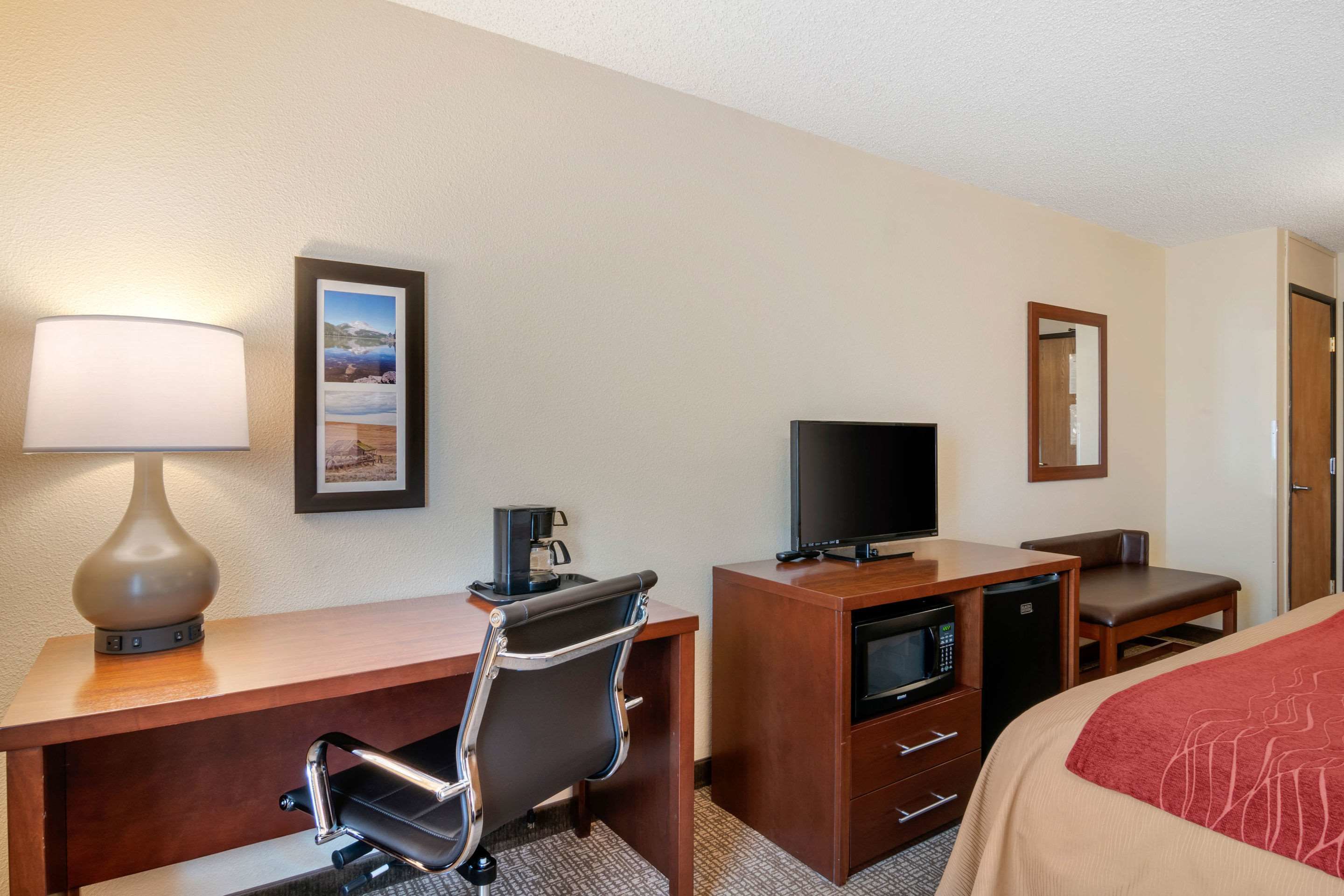 Habitación de hotel con un escritorio de madera, una silla de oficina negra, un televisor, un microondas, una cafetera, una cama con una colcha roja y un cuadro en la pared.