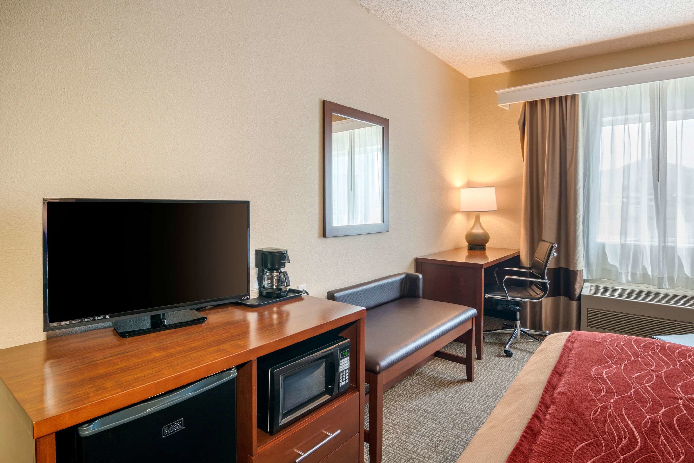 A hotel room with a flat-screen TV, a coffee maker, a microwave, a desk, a chair, a lamp, a bed, and a window with curtains.