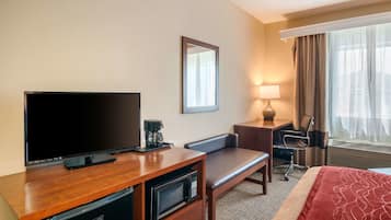 A hotel room with a flat-screen TV, a coffee maker, a microwave, a desk, a chair, a lamp, a bed, and a window with curtains.