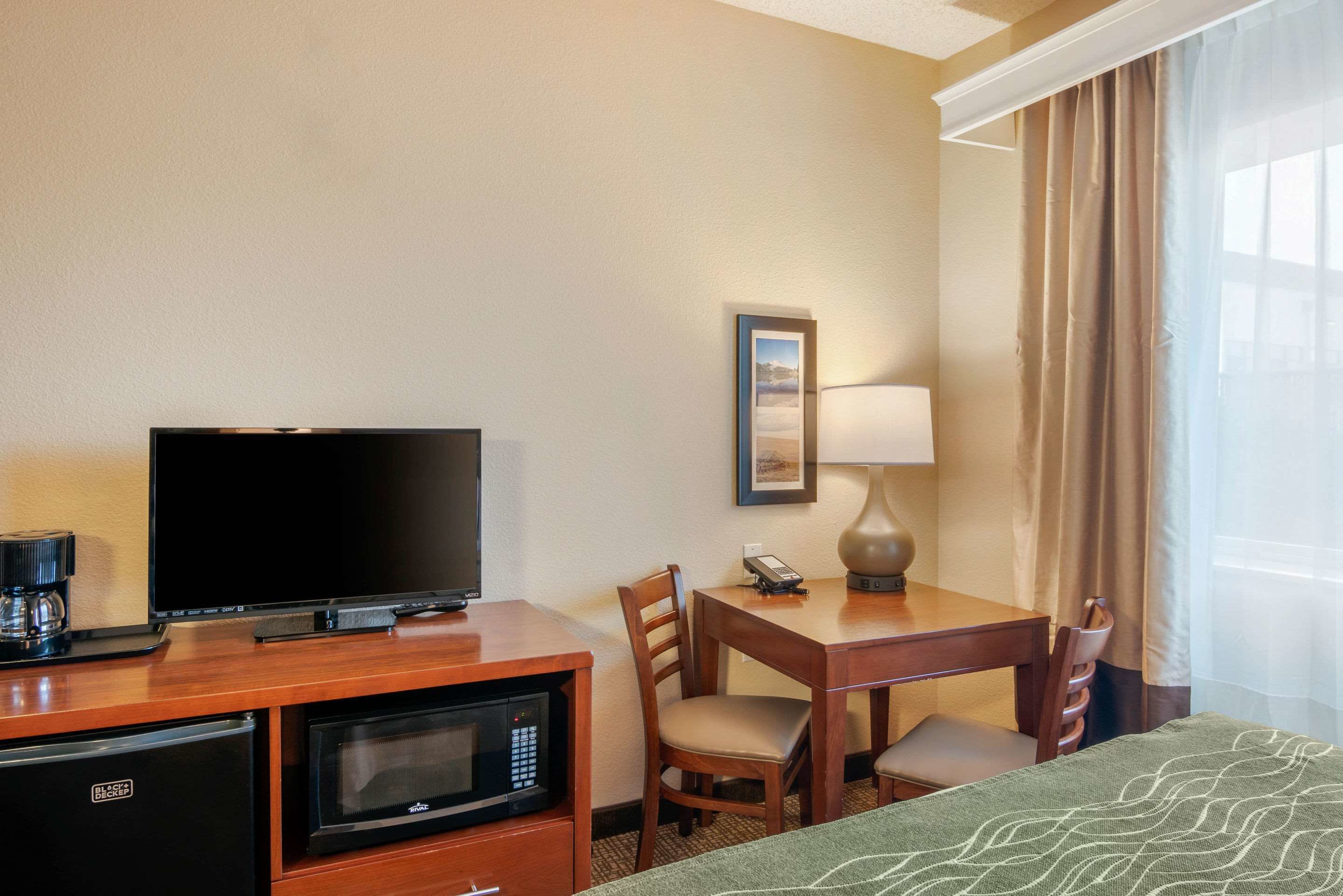 Quarto de hotel com televisão de tela plana, cafeteira, micro-ondas, mesa de jantar com cadeiras, abajur e cama com manta verde estampada.