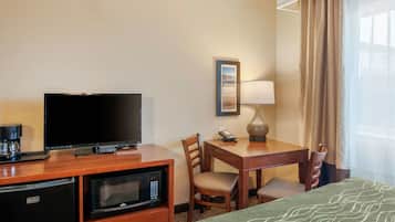 Quarto de hotel com televisão de tela plana, cafeteira, micro-ondas, mesa de jantar com cadeiras, abajur e cama com manta verde estampada.