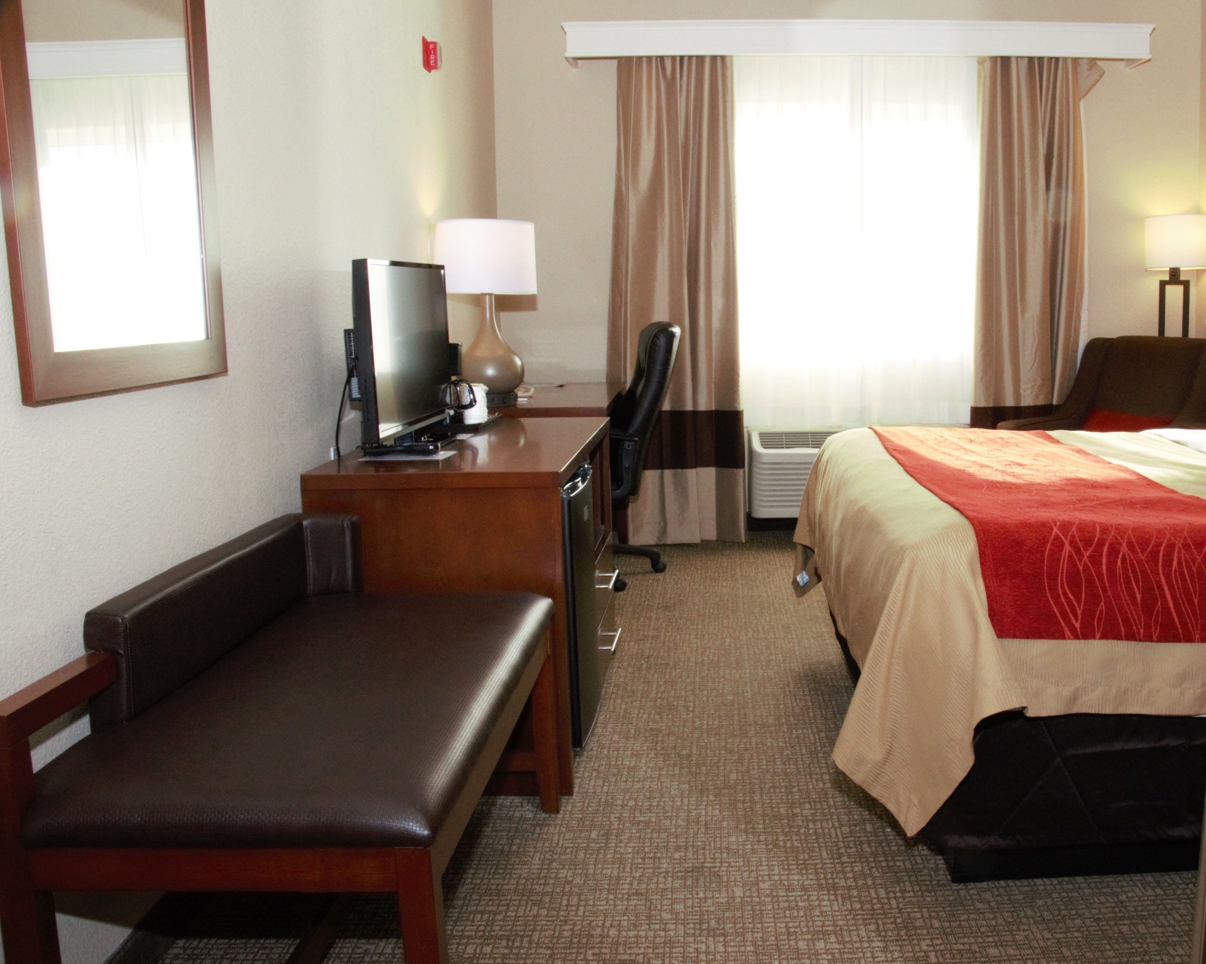 Una habitación de hotel con una cama, un escritorio con computadora y lámpara, una silla y un televisor.