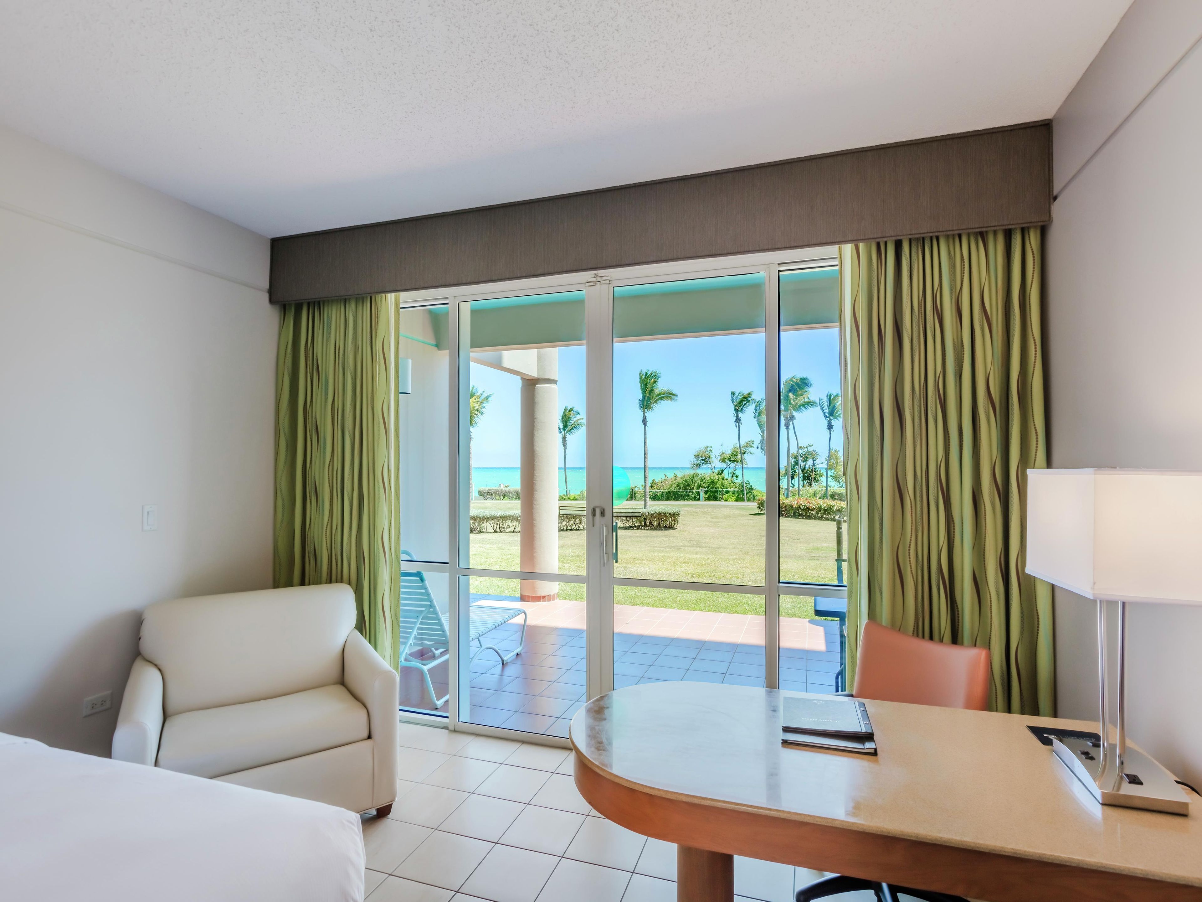 A hotel room with a bed, a chair, a table, and a view of the beach and palm trees through large glass doors.