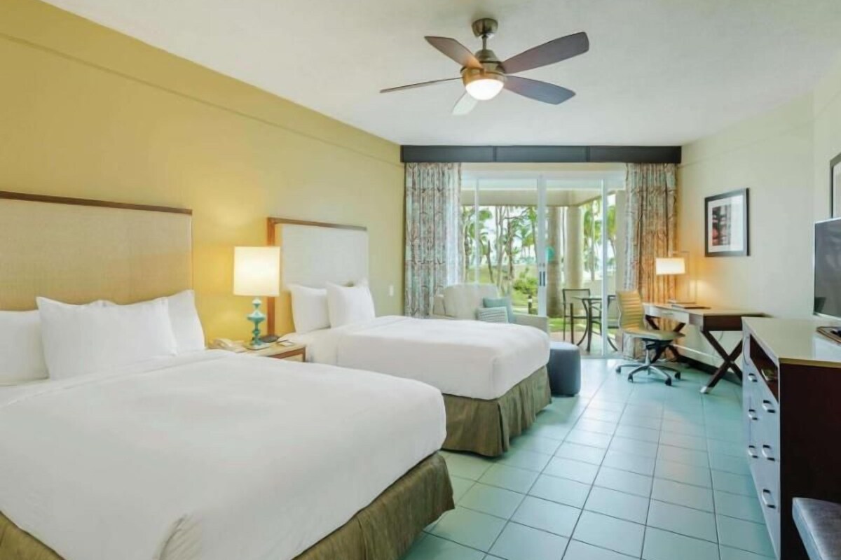 A hotel room with two beds, a desk, a chair, a television, and a ceiling fan.