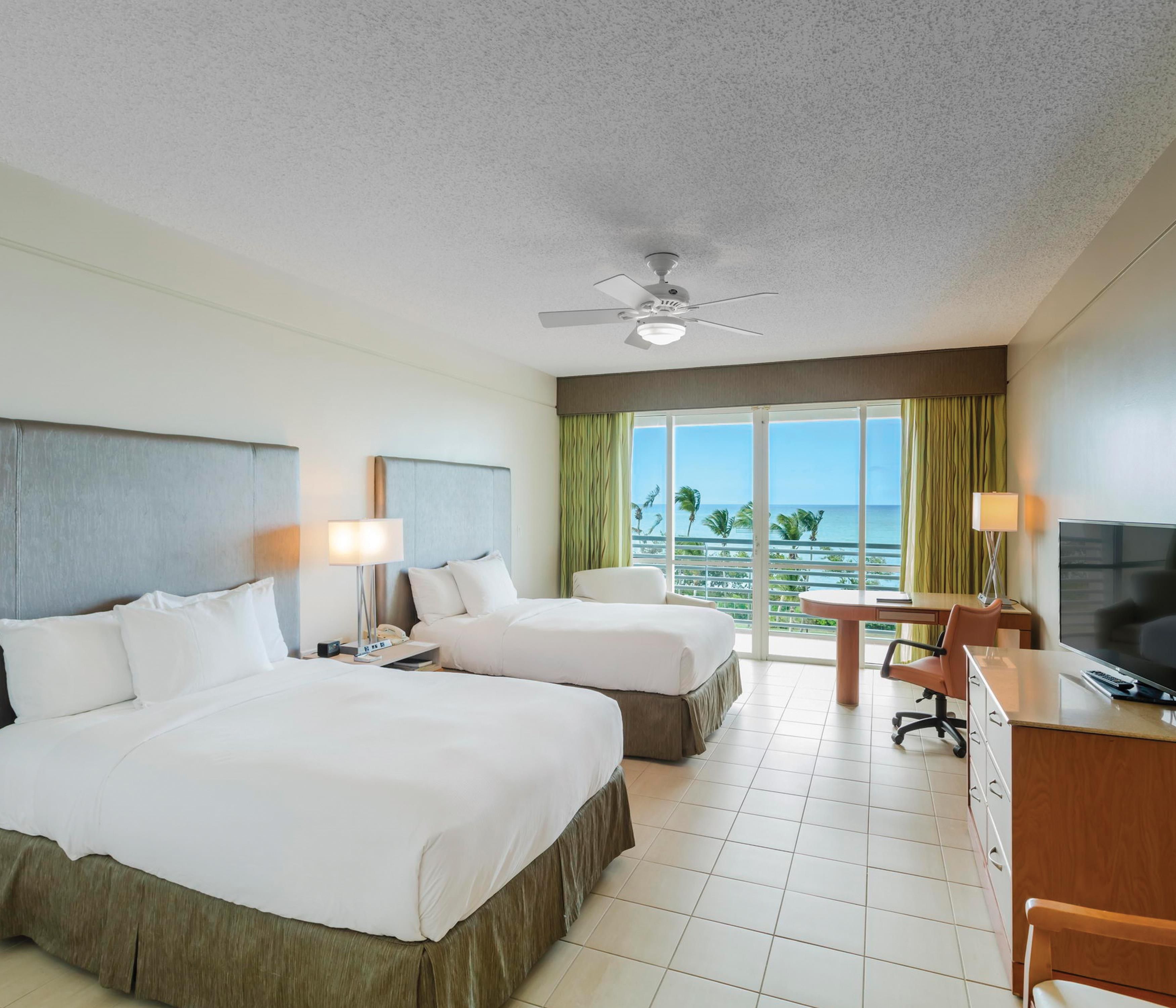 A hotel room with two beds, a desk with a chair, a television, and a balcony with a view of the ocean.