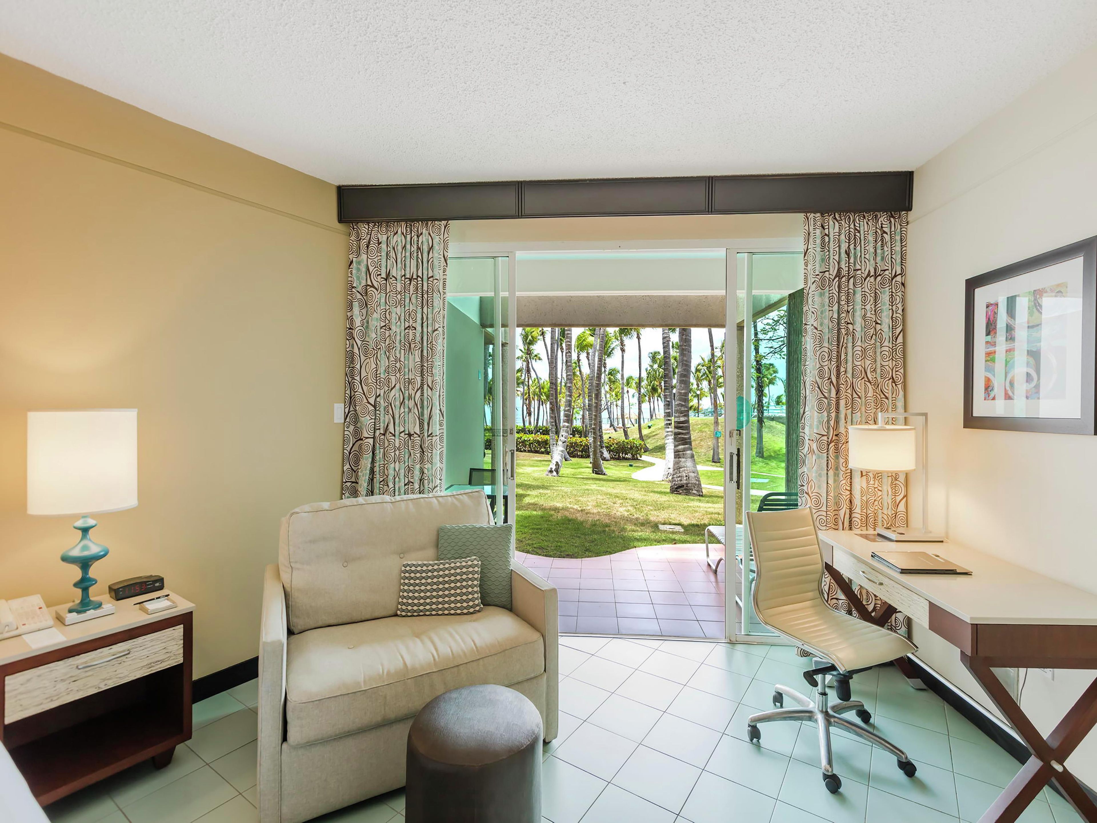 A hotel room with a sofa, a chair, a desk, a lamp, and a view of a garden with palm trees.