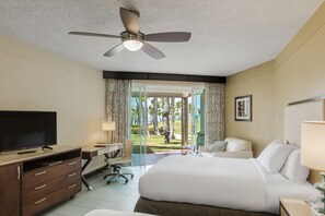 A hotel room with a bed, desk, TV, and a balcony with a view of palm trees.