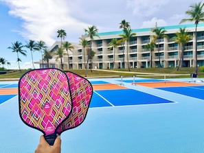 A hand holding a colorful paddle, a tennis court, and a building with multiple balconies and palm trees in the background.