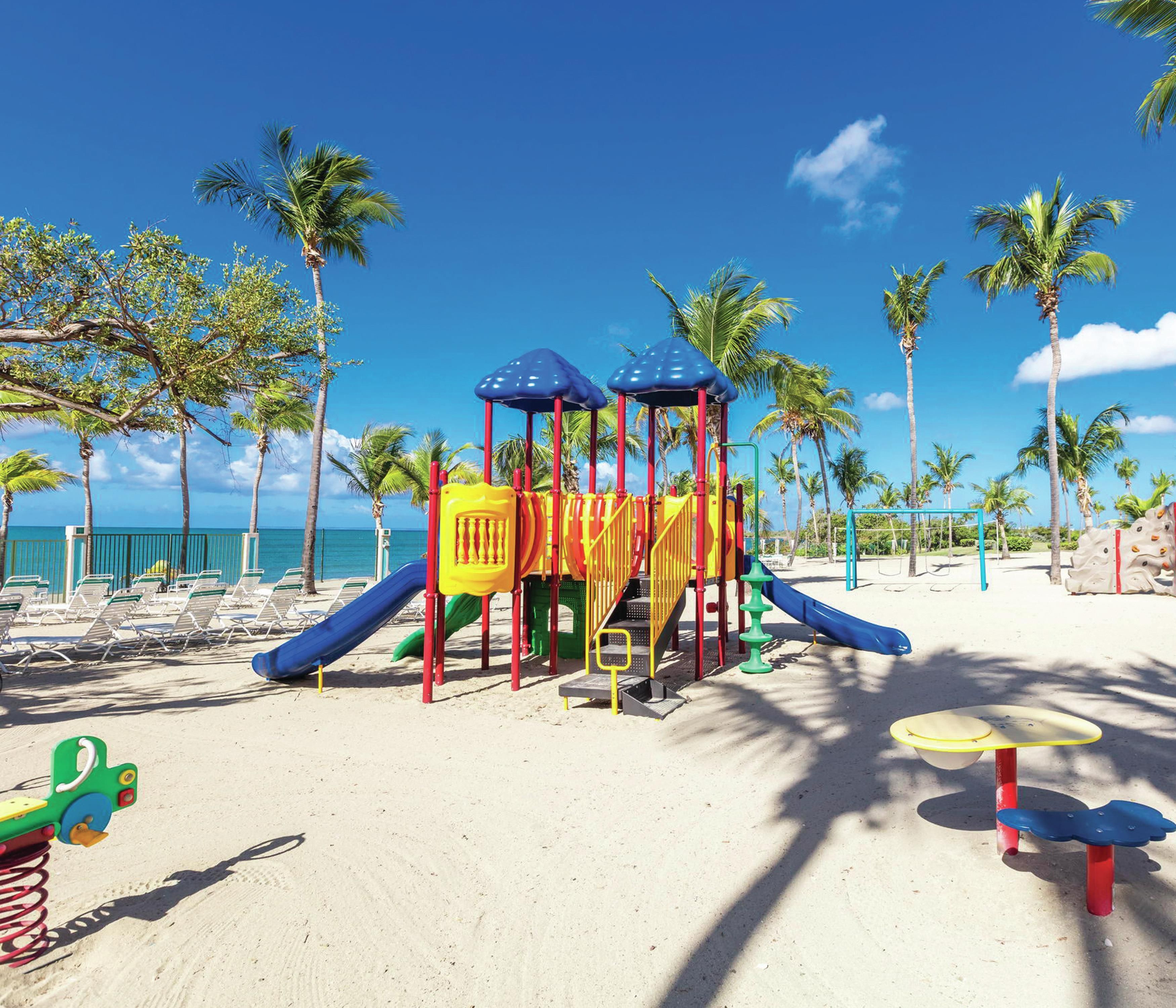 A beach playground with slides and climbing structures, surrounded by palm trees and beach chairs.