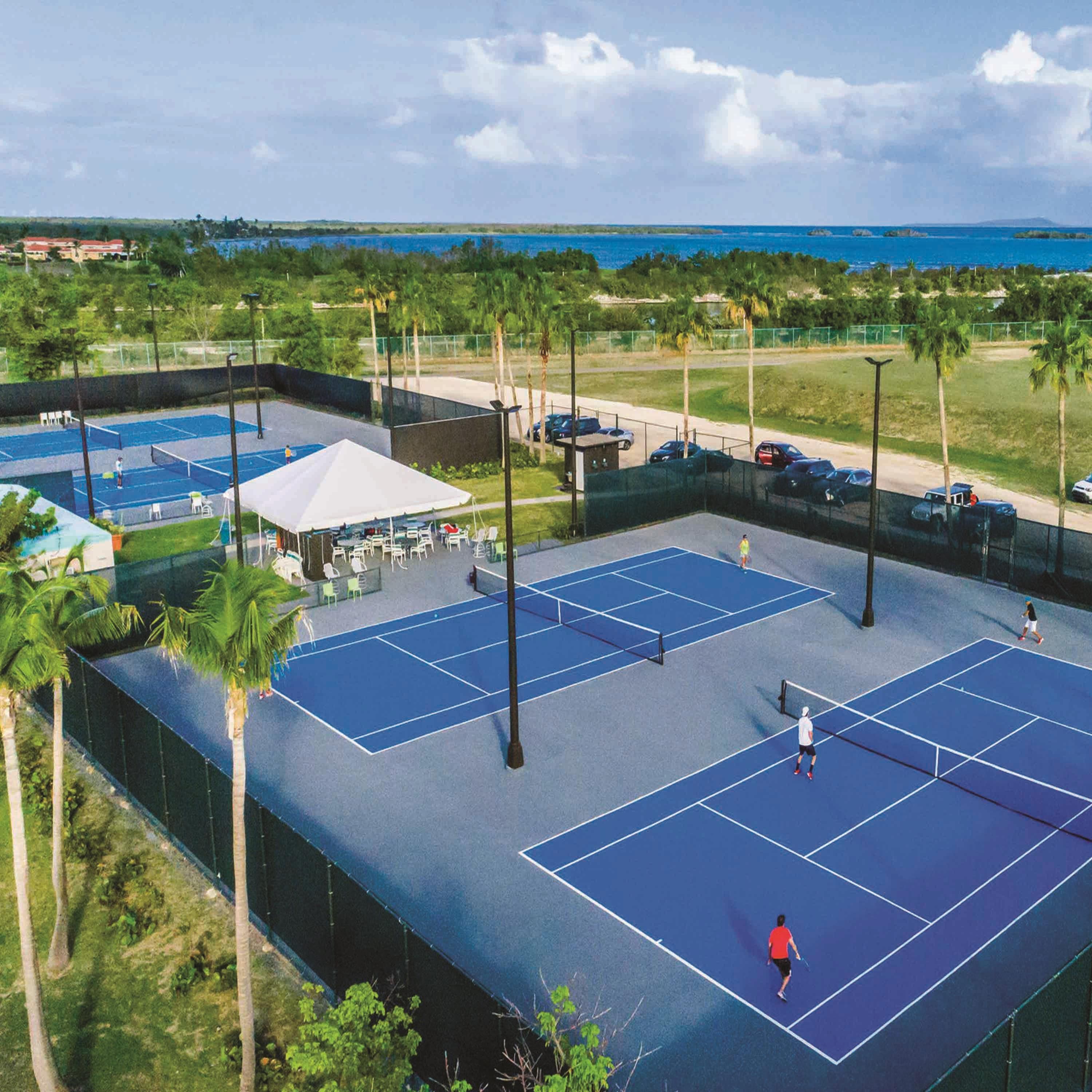 An outdoor tennis court with multiple courts, a covered seating area, and a view of the ocean.