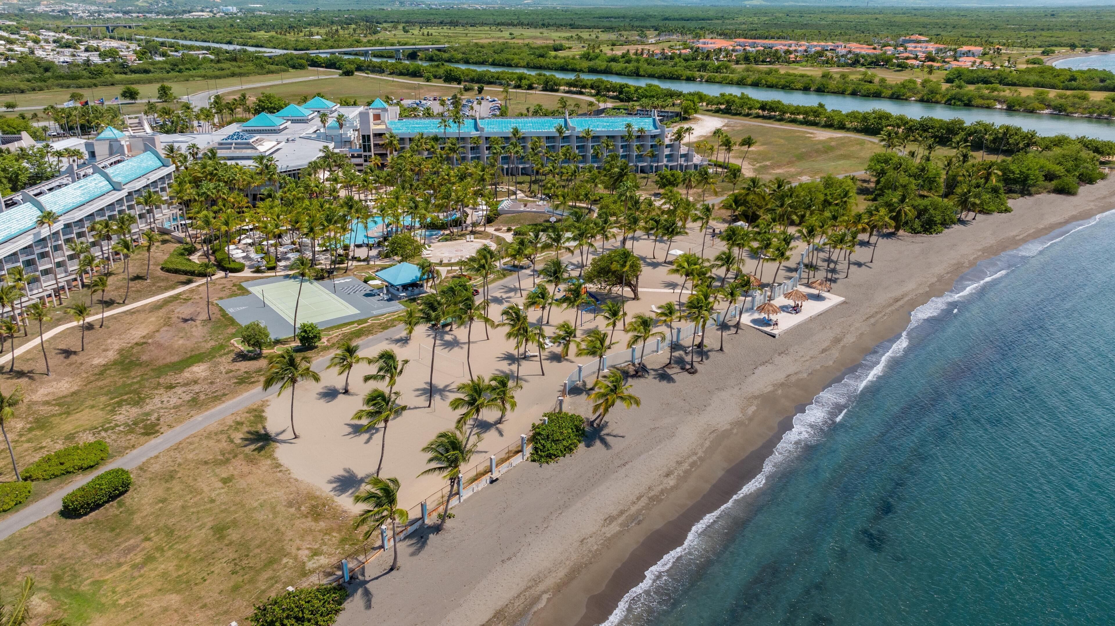 Luchtbeeld van een resort met een strand, palmbomen en een groot gebouw met een blauw dak.
