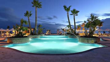 Une piscine bordée de palmiers et entourée de chaises longues, le tout situé au milieu d’une zone sablonneuse.