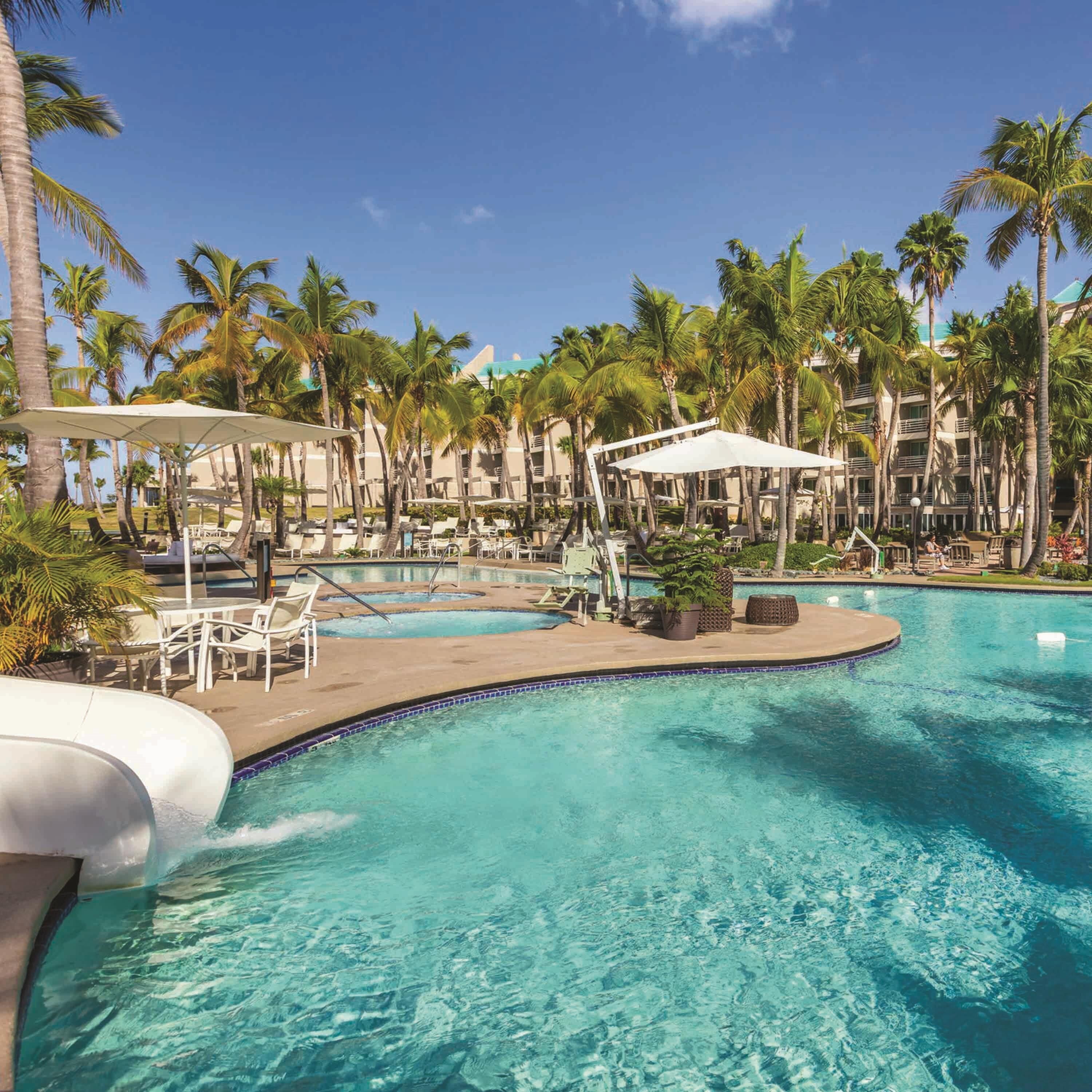 Ein Poolbereich eines Resorts mit mehreren Liegestühlen und Sonnenschirmen, umgeben von Palmen.