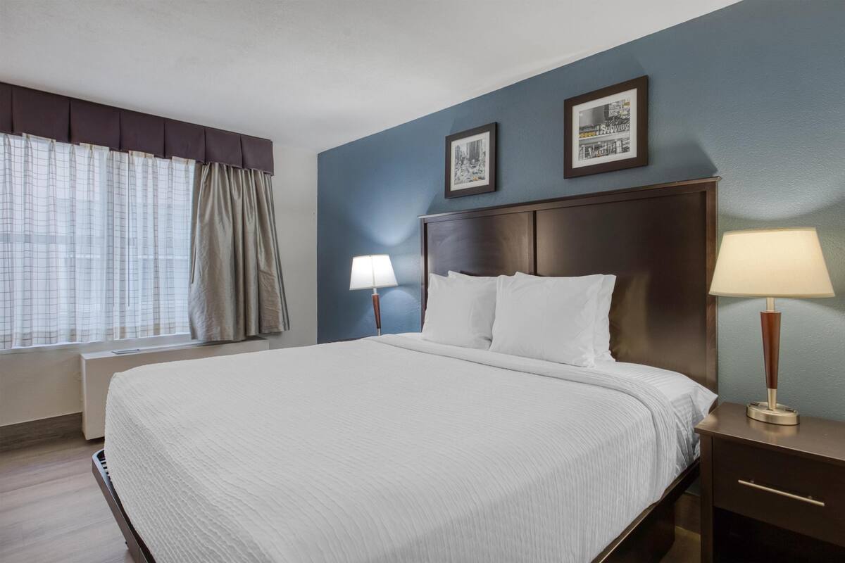 A hotel room with a large bed, two bedside tables with lamps, a window with curtains, and two framed pictures on the wall.