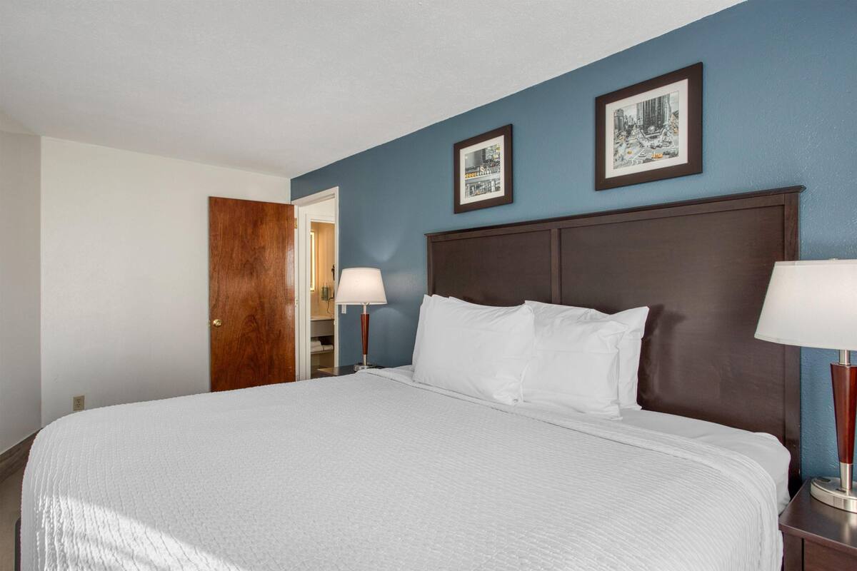 A hotel room with a large bed, two bedside lamps, a wooden door, and two framed pictures on the wall.