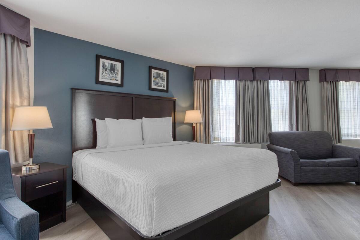A hotel room with a large bed, two bedside tables with lamps, a grey armchair, and two framed pictures on the wall.