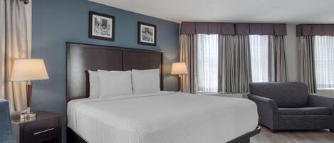 A hotel room with a large bed, two bedside tables with lamps, a grey armchair, and two framed pictures on the wall.