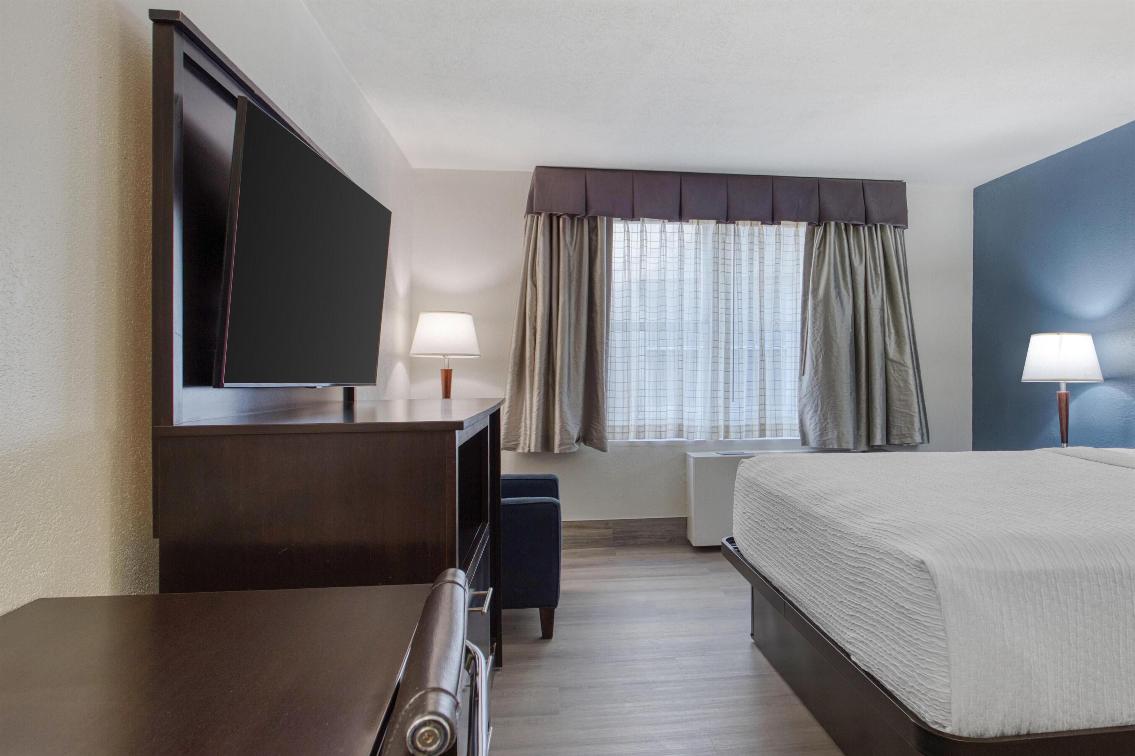 A hotel room with a bed, a desk with a flat-screen TV, a bedside table with a lamp, a window with curtains, and a wall-mounted air conditioning unit.