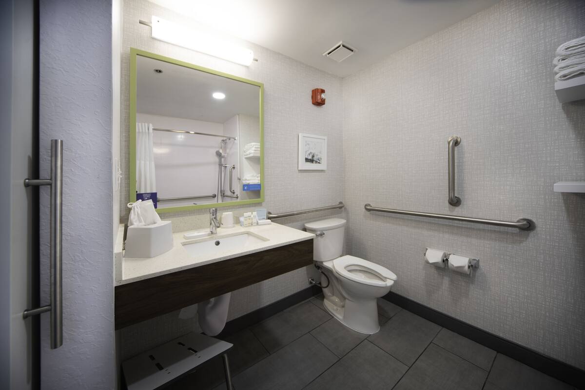 Un baño moderno con lavamanos, espejo y retrete blancos. Incluye un pasamanos para accesibilidad y un perchero.